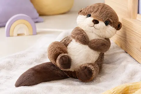 Peluche loutre douce avec ventre animé et queue satinée, posée dans un décor clair aux tons lilas et crème