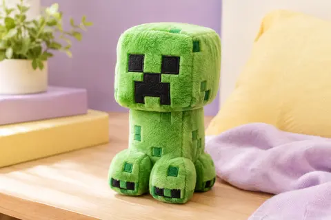 Peluche pixelisée inspirée d’un Creeper de Minecraft, posée dans un décor doux lilas et crème.