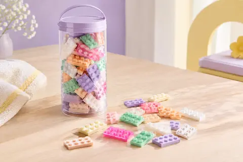 Briques de construction nacrées aux tons pastel disposées près de leur tube de rangement sur du bois clair.