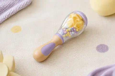 Hochet maracas en plastique avec billes multicolores et poignée, posé sur un fond crème aux touches lilas.