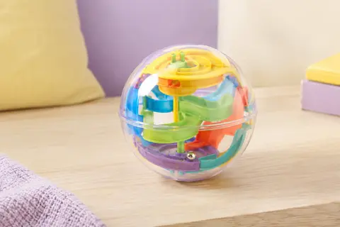 Sphère de labyrinthe 3D Perplexus Rebel multicolore posée sur une table en bois clair dans un décor lilas.