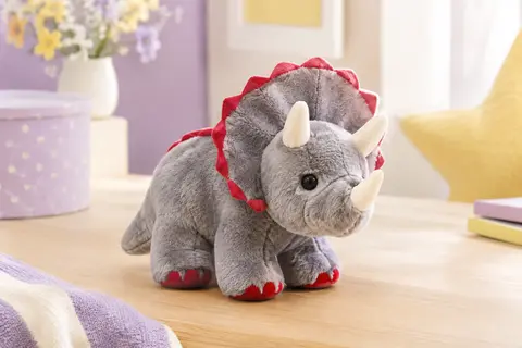 Peluche tricératops douce aux détails rouges et gris posée sur du bois clair dans un décor pastel